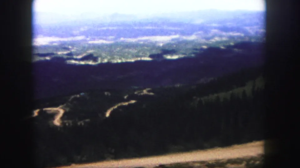1958 Colorado Springs home-movie frame looking from a high mountain vantage across forested hills, a winding road, and distant haze.