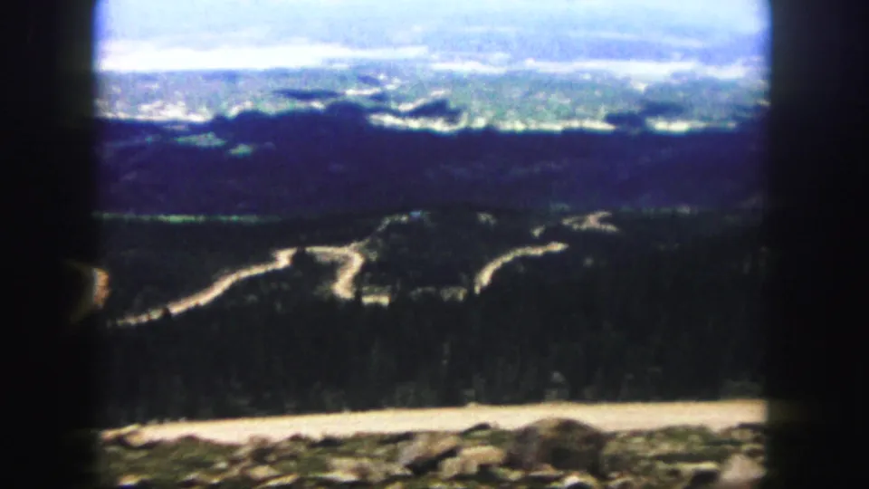 High-vantage 1958 Colorado Springs frame showing evergreen-covered hills, serpentine roads, and a distant hazy valley.