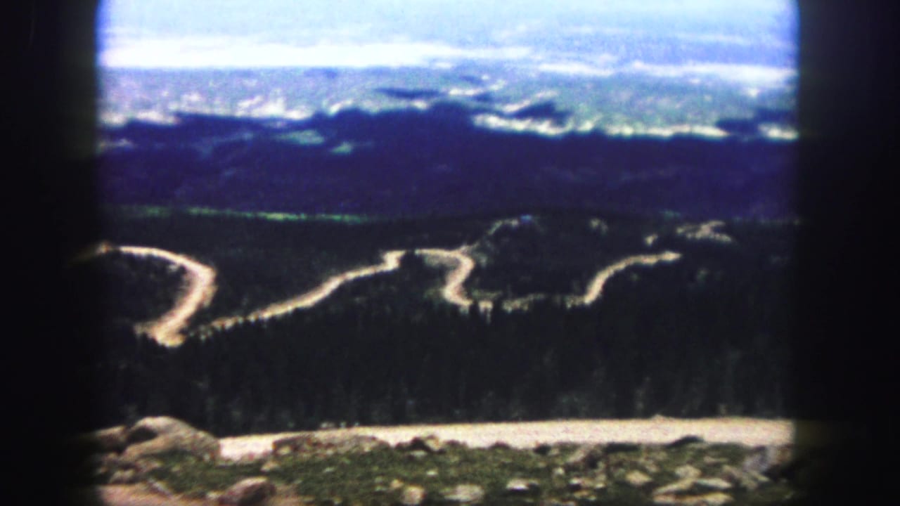 1958 Colorado Springs home-movie frame showing a winding mountain road through forested hills, seen from a high viewpoint with dark film borders.