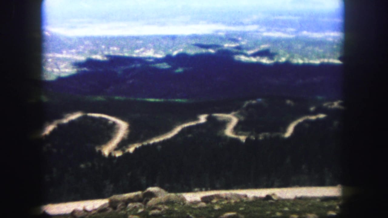 A 1958 Colorado Springs 8mm view of a sharply curving mountain road below shadowed forested slopes and a hazy valley horizon.
