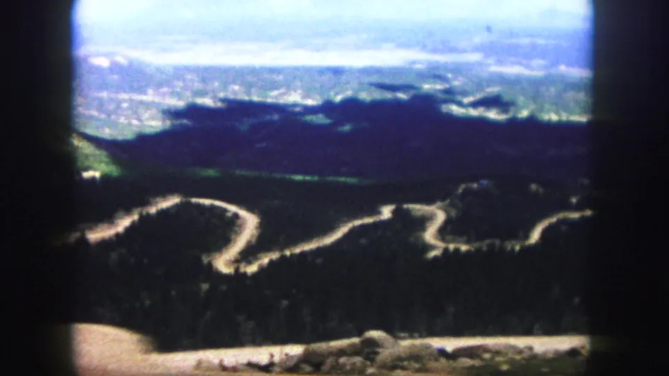 Soft 8mm frame of a winding road through wooded Colorado Springs hills, with bright sky, distant haze, and visible black projection borders.