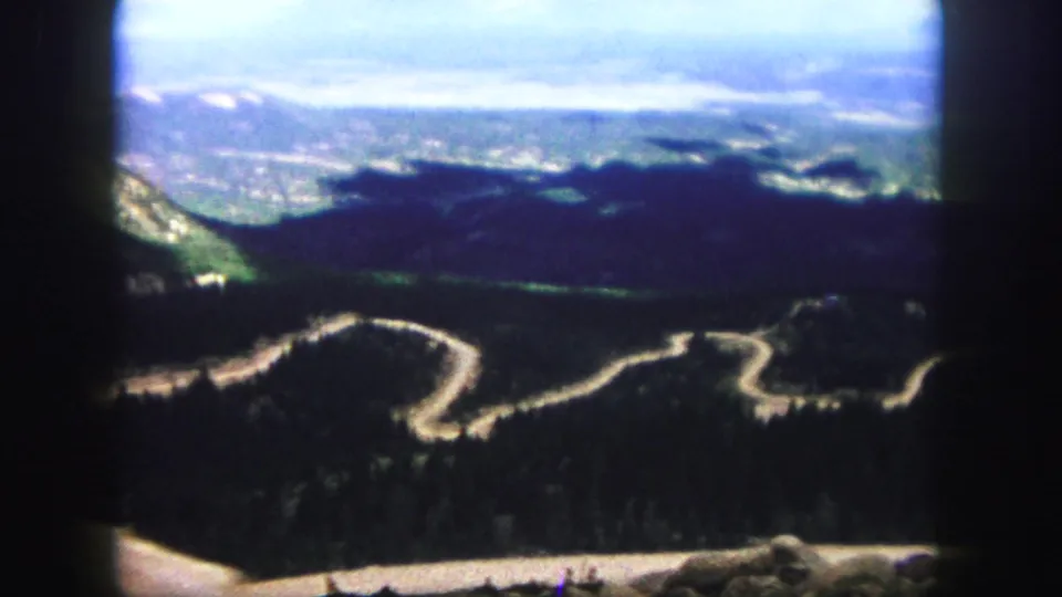 Colorado Springs 1958 home-movie still showing a pale mountain road winding through dense forest, with distant terrain and dark 8mm frame edges.