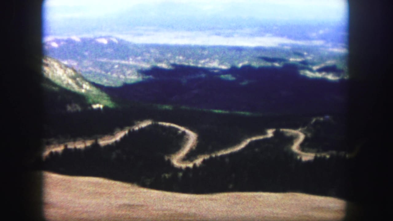 1958 8mm home-movie view near Colorado Springs showing a winding mountain road, forested slopes, distant ridges, and dark projected frame edges.