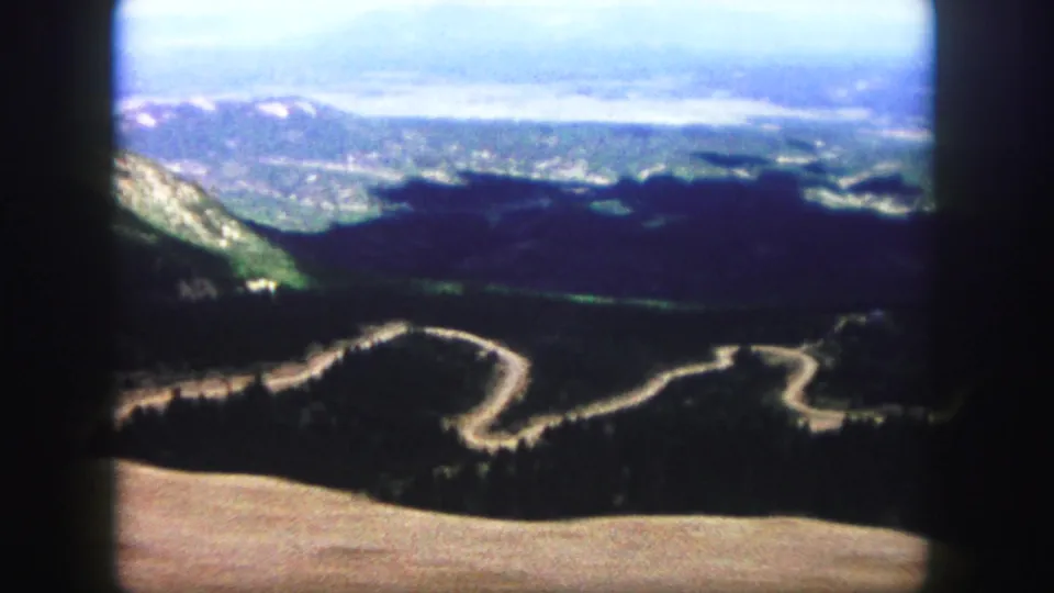 1958 8mm home-movie view near Colorado Springs showing a winding mountain road, forested slopes, distant ridges, and dark projected frame edges.