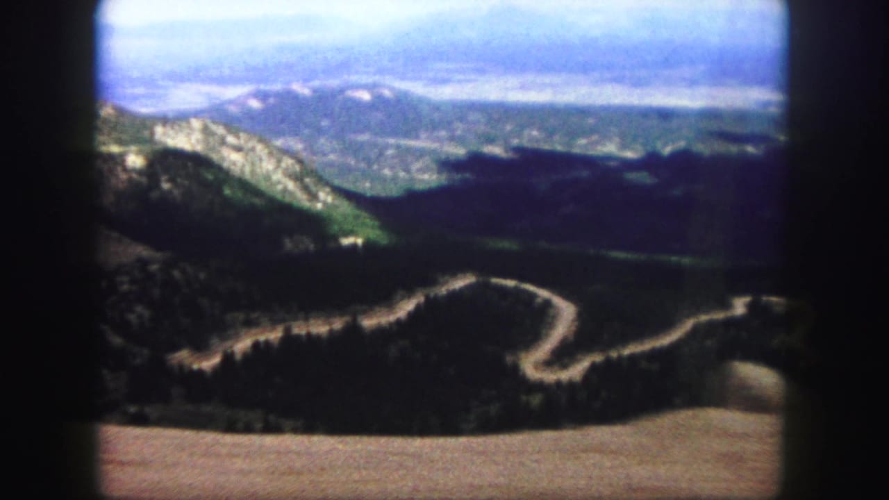 Similar elevated mountain view with a pale road curving across dark wooded terrain.