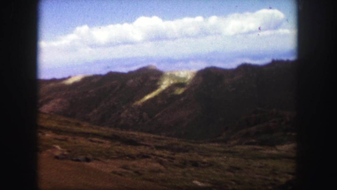 1958 Colorado Springs home-movie frame showing rugged mountain ridges, pale slope patches, open foreground, and clouded blue sky.