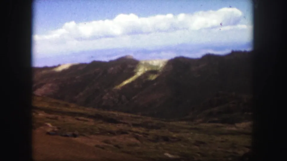 1958 Colorado Springs home-movie frame showing rugged mountain ridges, pale slope patches, open foreground, and clouded blue sky.