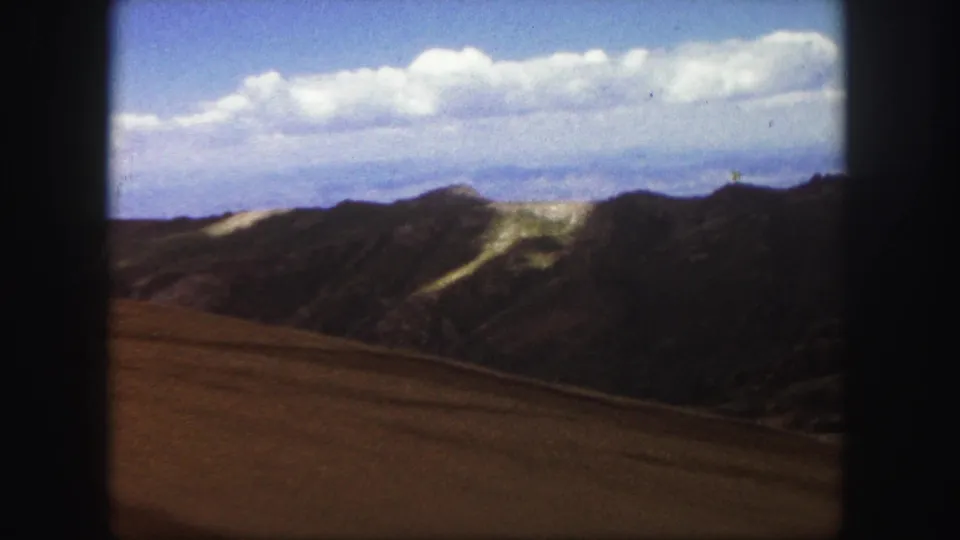 1958 Colorado Springs home-movie frame with brown open ground, dark mountain ridges, pale slope marks, and a bright cloud band.