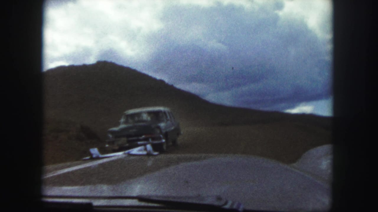 1958 Colorado Springs home-movie frame looking through a car windshield as a dark automobile approaches on a winding road beneath a cloudy sky.