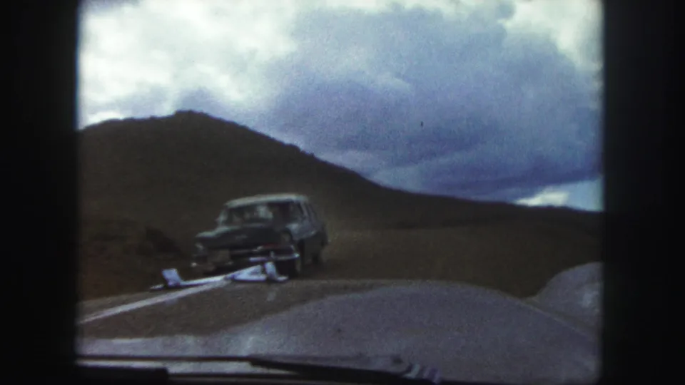 1958 Colorado Springs home-movie frame looking through a car windshield as a dark automobile approaches on a winding road beneath a cloudy sky.