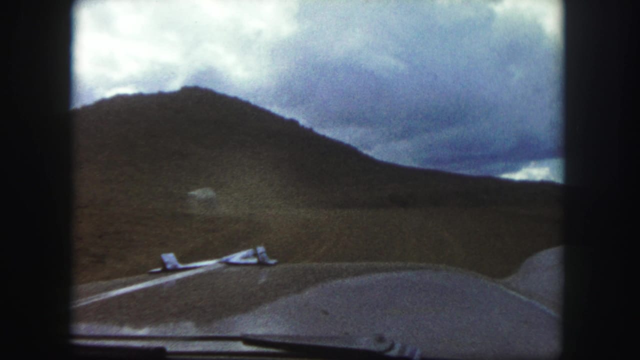 Windshield view of a road curving past a hillside under heavy clouds.