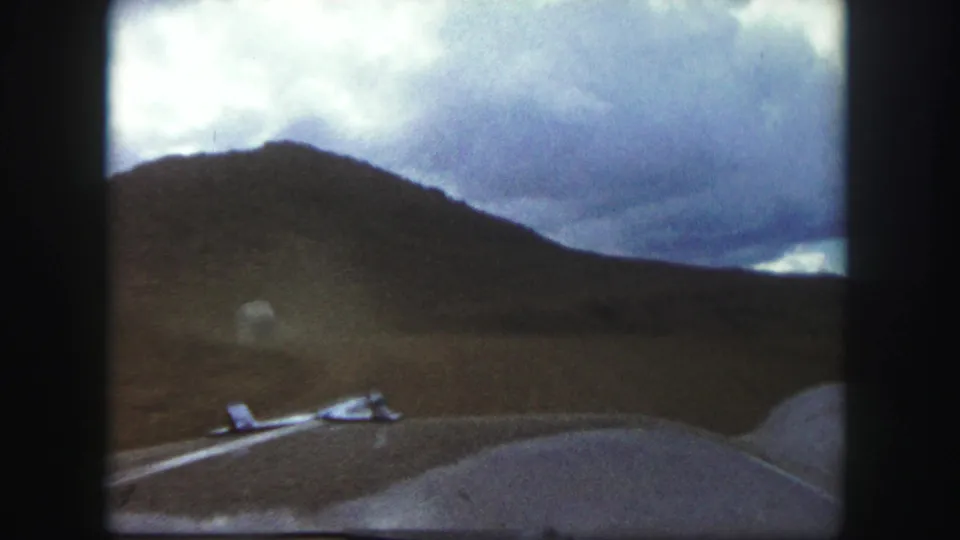 Driver’s-eye 1958 Colorado Springs view through the windshield toward a low road, open hillside, and overcast sky.