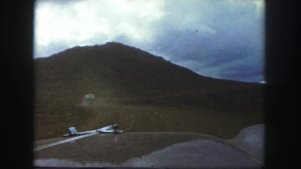 Final 1958 Colorado Springs windshield frame showing the road ahead, rounded hood, open hillside, and heavy clouds in a grainy 8mm view.