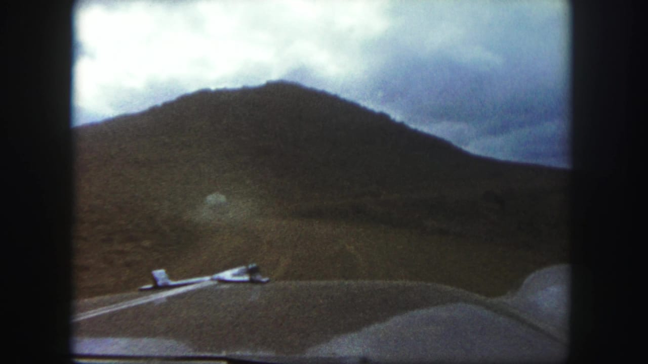 1958 Colorado Springs home-movie view from a car hood, looking down an unpaved road toward a barren hillside beneath clouds.