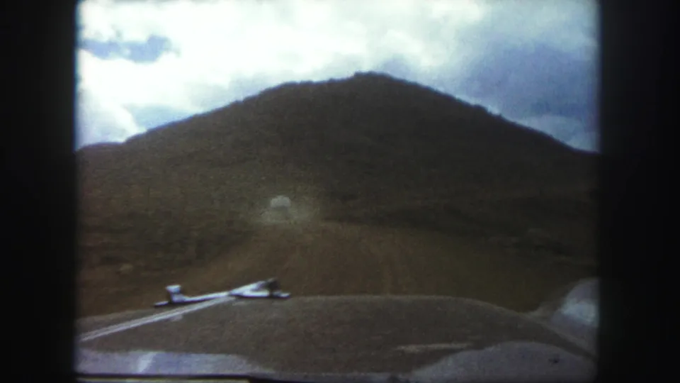 Car-hood perspective near Colorado Springs in 1958, showing a rough road leading toward a scrubby hill under bright clouds.