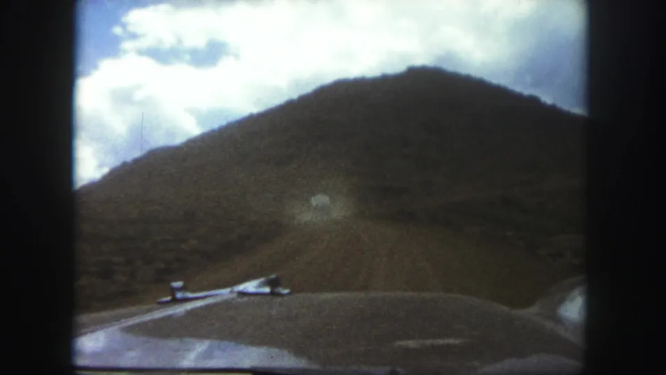 8mm home-movie frame from a moving car near Colorado Springs, with the dirt road rising toward a dark hillside and cloudy sky.