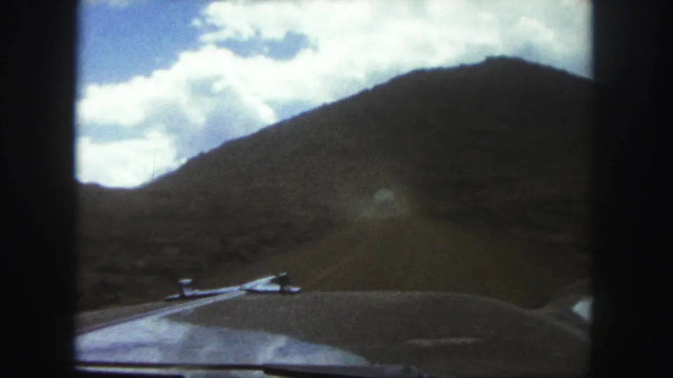 Late-1950s car-hood view in Colorado Springs, showing an unpaved road, open scrubland, and a dark hill under a partly cloudy sky.