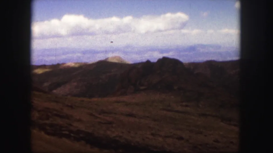 1958 Colorado Springs 8mm view of a dark rocky ridge, dry foreground, distant mountains, and scattered clouds.