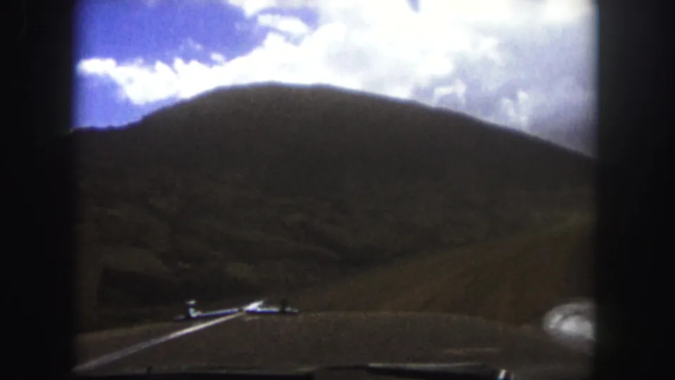 Vintage 8mm Colorado road scene with a small car ahead, curving pavement, hills, and bright sky.
