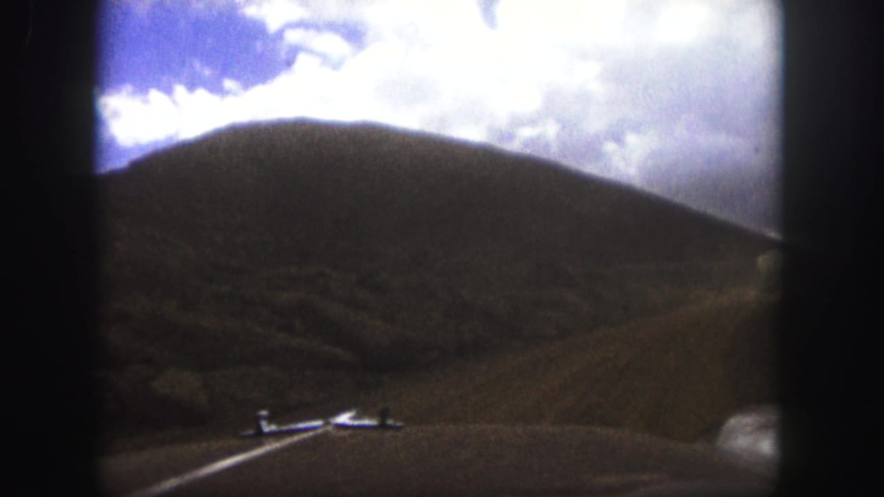 View over a car hood toward a dusty road curving through brown hills under bright clouds.