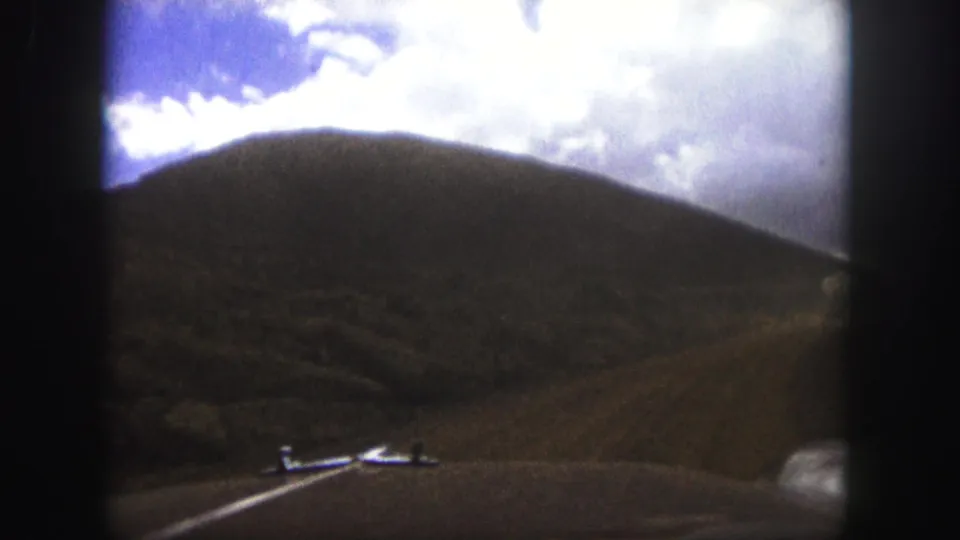 1958 Colorado Springs home-movie view from inside a car, looking over the hood at an unpaved road and rounded hillside beneath a cloudy sky.