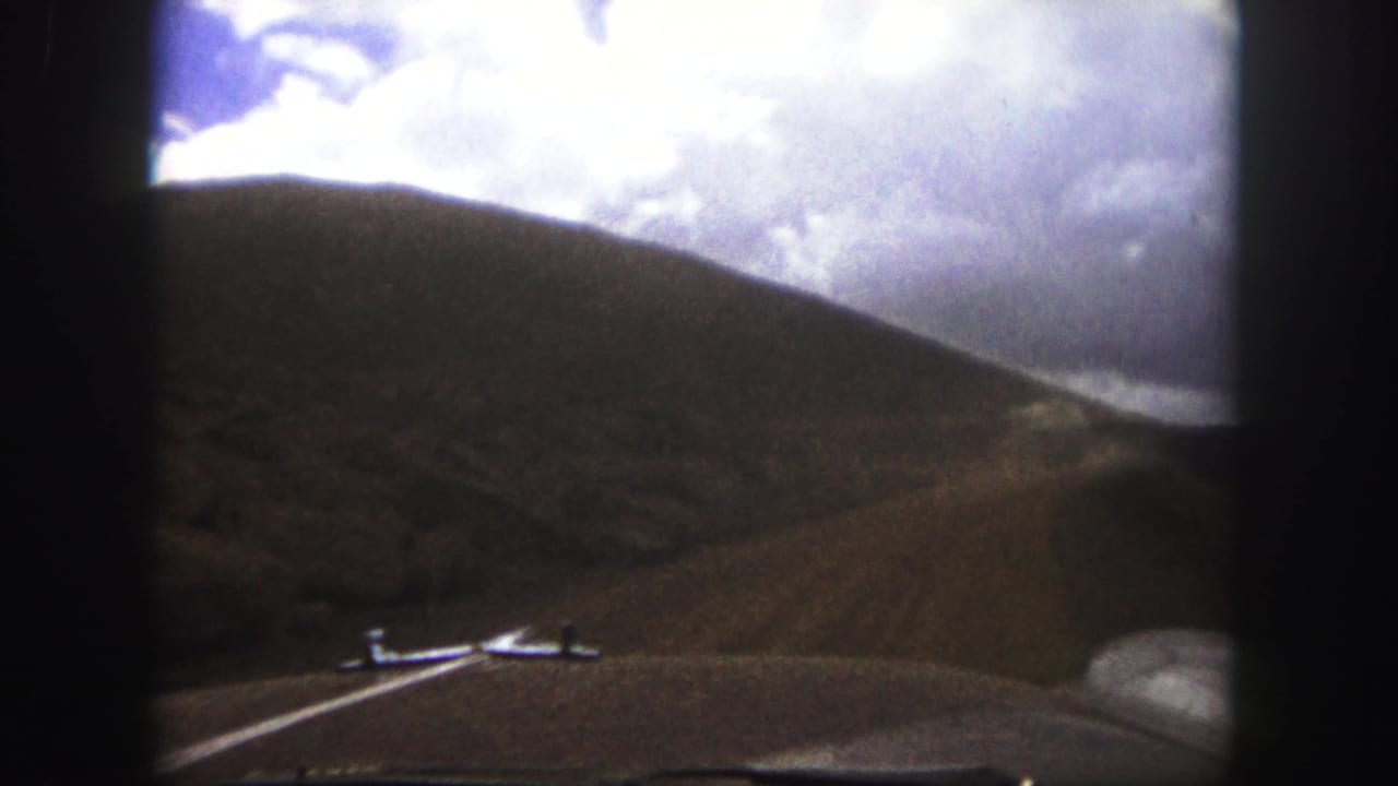 Car hood foreground with an unpaved road rising between hills beneath bright clouds.