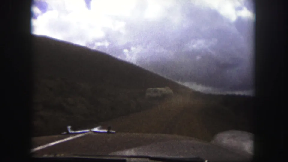 Late-frame 1958 Colorado Springs driving view with the car hood at bottom, a dusty road ahead, and open hills under heavy clouds.