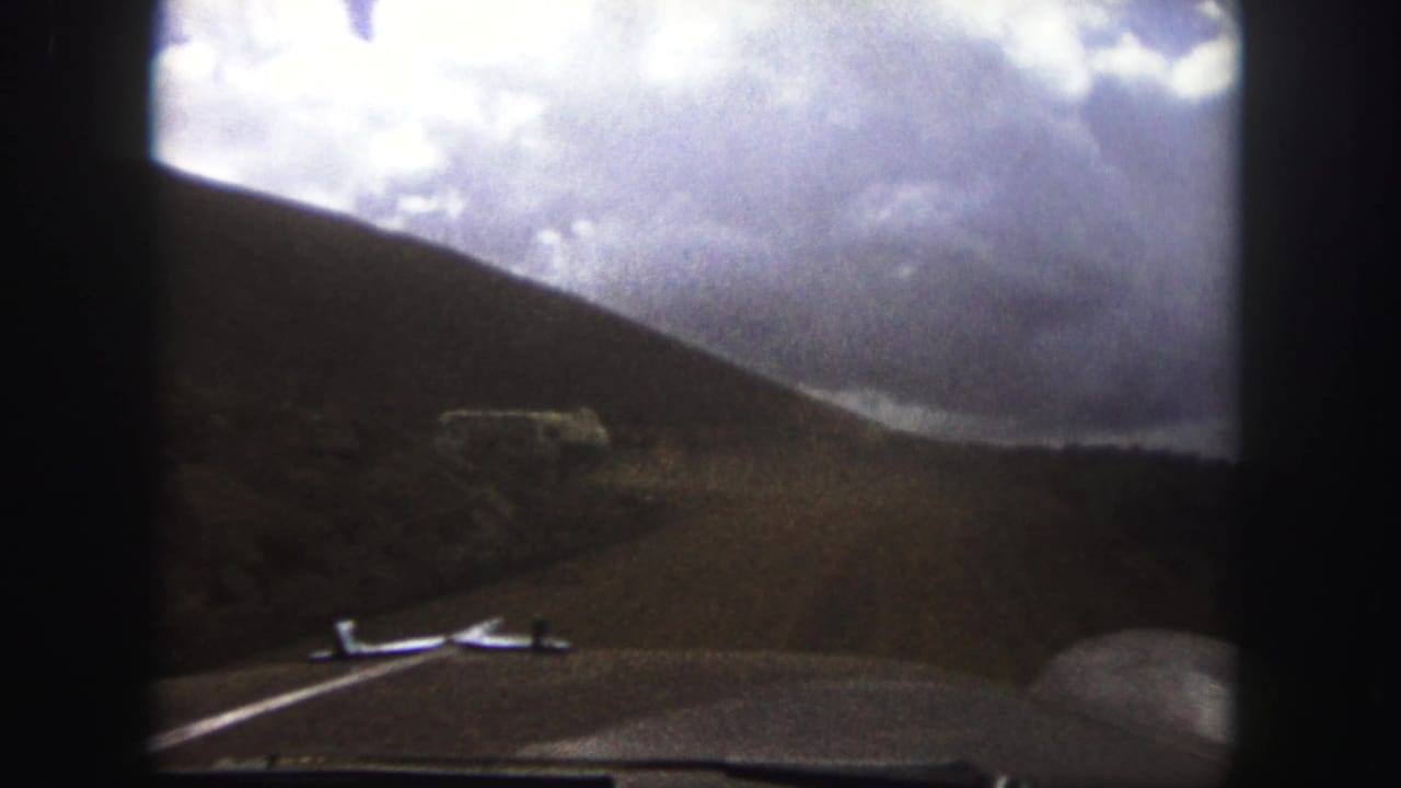 Windshield view of a curving dirt road, hillside, and white bus beneath a dramatic cloud bank.