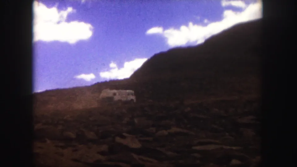 Colorado Springs 1958 8mm frame with a white bus moving through rocky mountain terrain under a brighter blue sky.