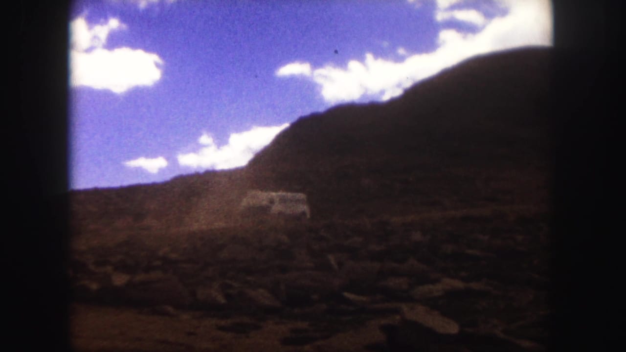 1958 Colorado Springs 8mm frame showing a rocky dirt road, mountain slope, bright sky, and a red truck approaching from the right edge.