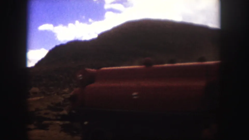 1958 Colorado Springs 8mm frame showing the side of a red truck as it passes on a rough road beneath a dark mountain slope.