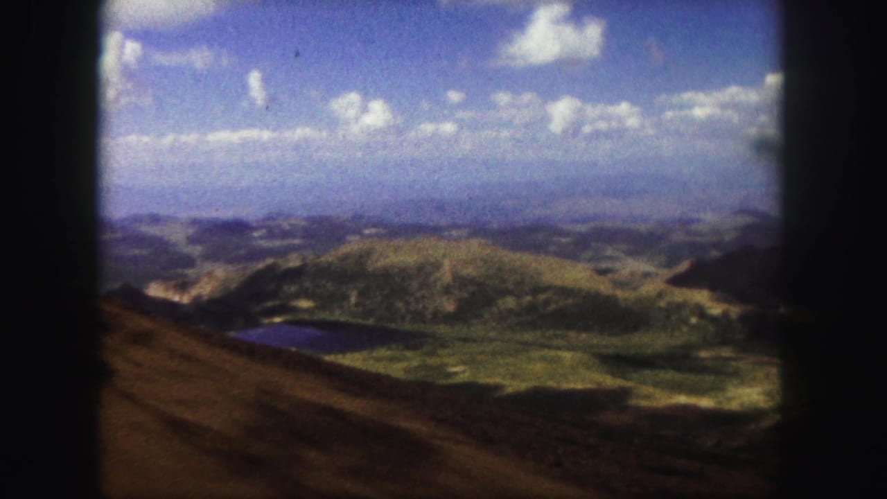 Rolling hills and a small lake seen from a high roadside overlook under a blue sky.