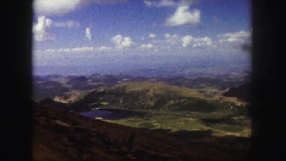 1958 Colorado Springs home-movie still showing a small lake, green hills, rocky foreground, and bright clouds from a roadside overlook.