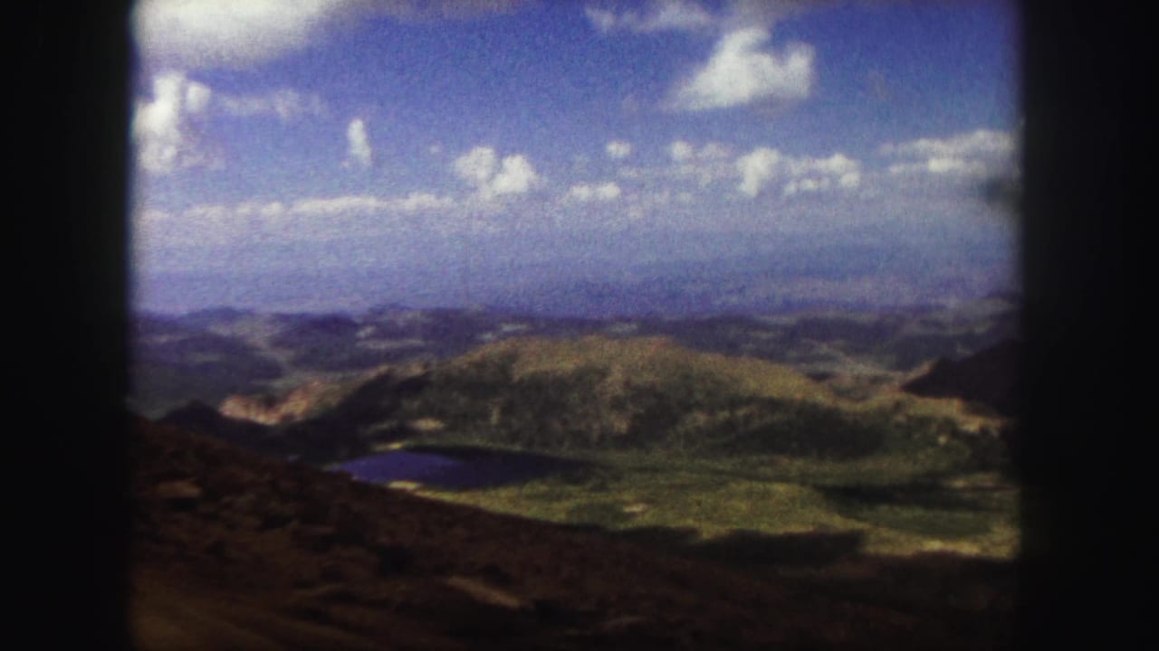 1958 Colorado Springs 8mm frame looking across green hills, a small lake, distant valleys, and a clouded blue sky.