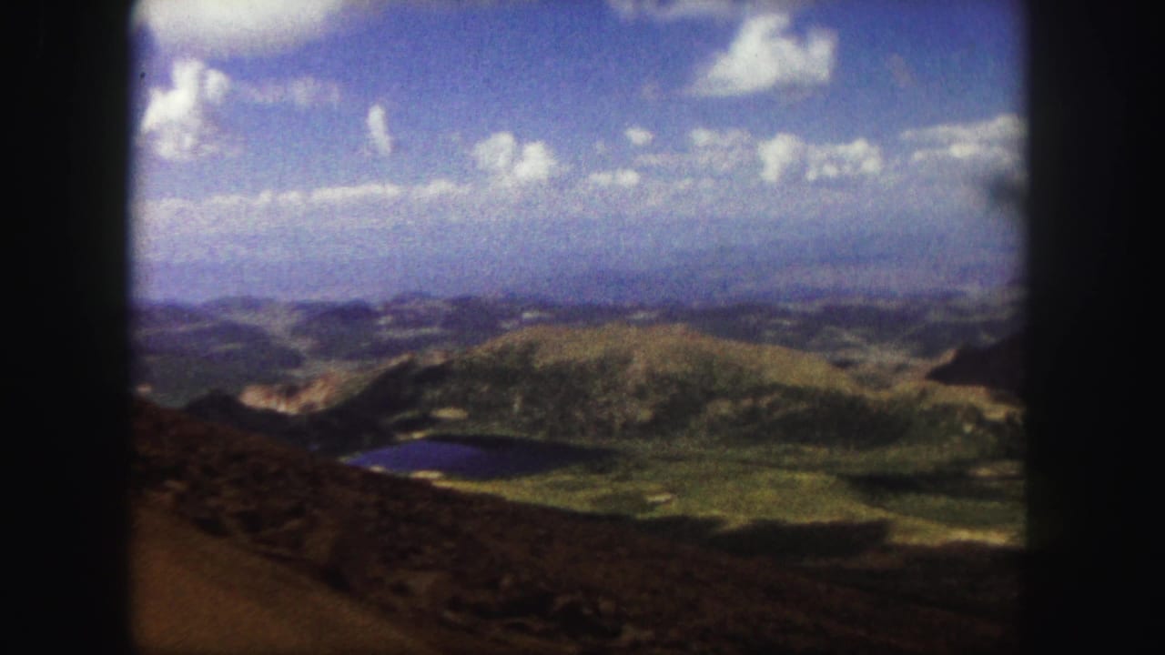 Mountain panorama with rounded hills, valley floor, small lake, and bright clouds.