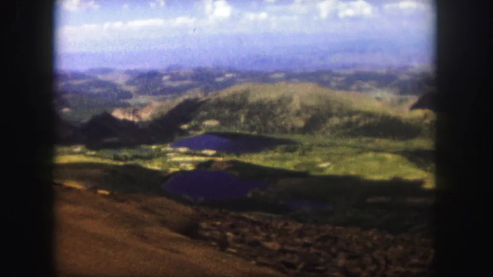 Colorado Springs 1958 home-movie frame showing a wide mountain landscape, dark lake, green hills, and hazy horizon.