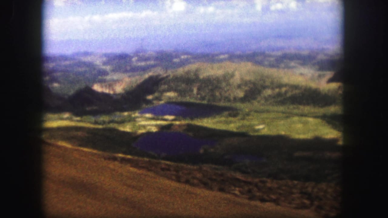 1958 Colorado Springs 8mm frame looking from a brown ridge across green hills, dark lakes, and a hazy valley under blue sky.