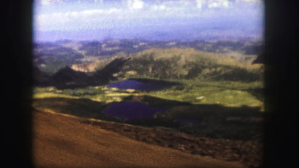 1958 Colorado Springs 8mm frame looking from a brown ridge across green hills, dark lakes, and a hazy valley under blue sky.