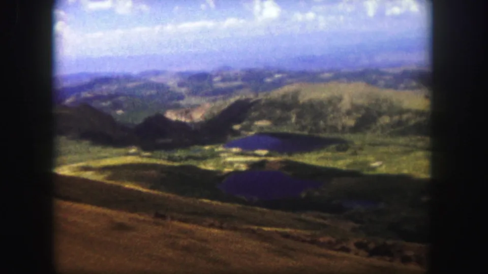 A soft, grainy 1958 Colorado Springs 8mm view across a ridge foreground, dark lakes, green hills, and hazy sky.