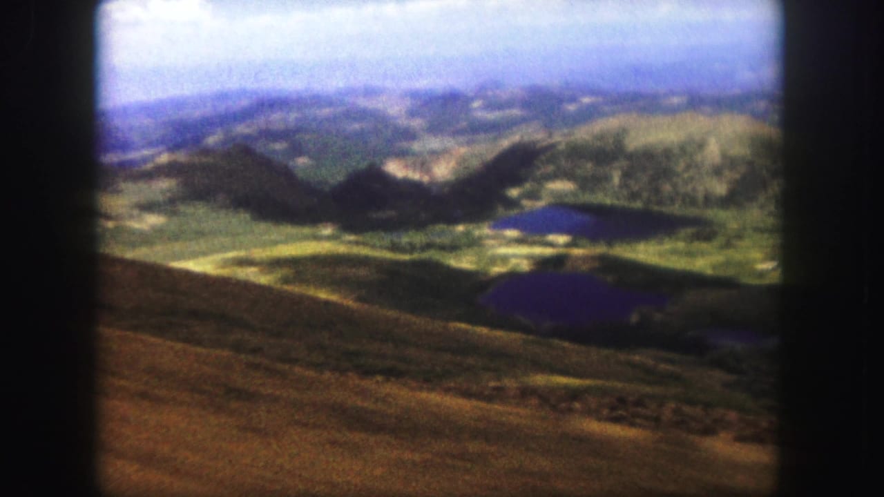 1958 Colorado Springs-area home-movie frame showing a hazy mountain landscape with small blue-purple lakes beyond a sunlit brown slope.