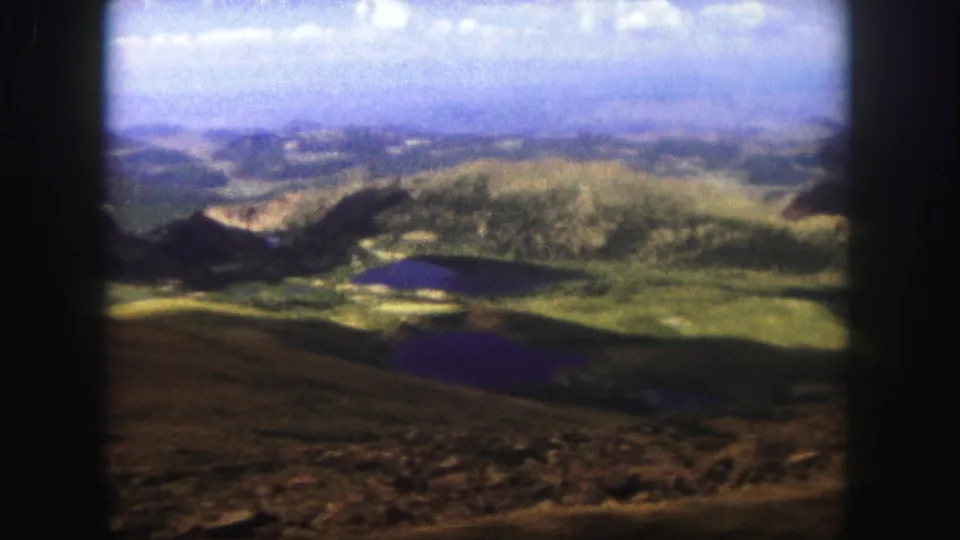 Wide 1958 Colorado Springs-area scenic frame with layered ridges, small lakes, green terrain, and the visible edge of the 8mm film scan.