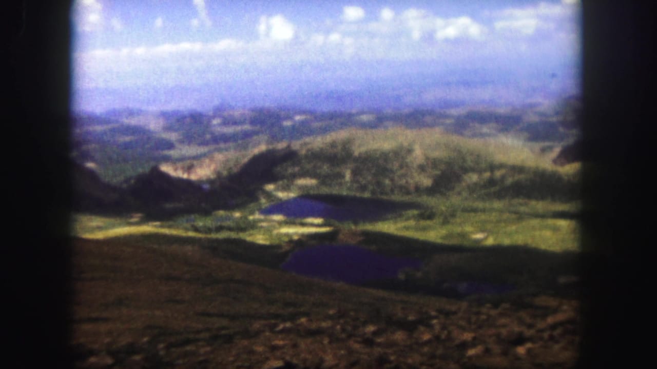 1958 Colorado Springs 8mm frame showing a mountain overlook across rocky foreground, green valley slopes, dark lakes, and distant hazy ridges.