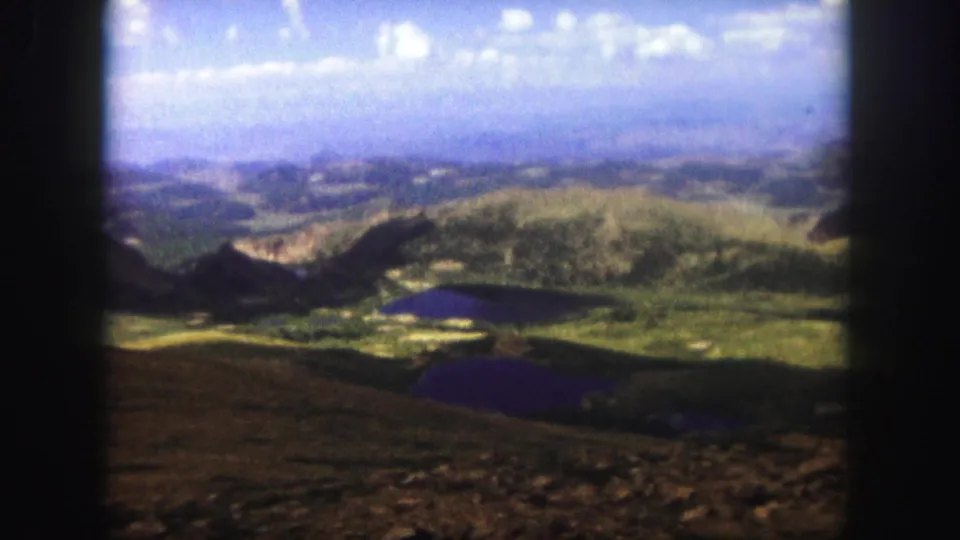 1958 Colorado Springs 8mm frame showing a mountain overlook across rocky foreground, green valley slopes, dark lakes, and distant hazy ridges.