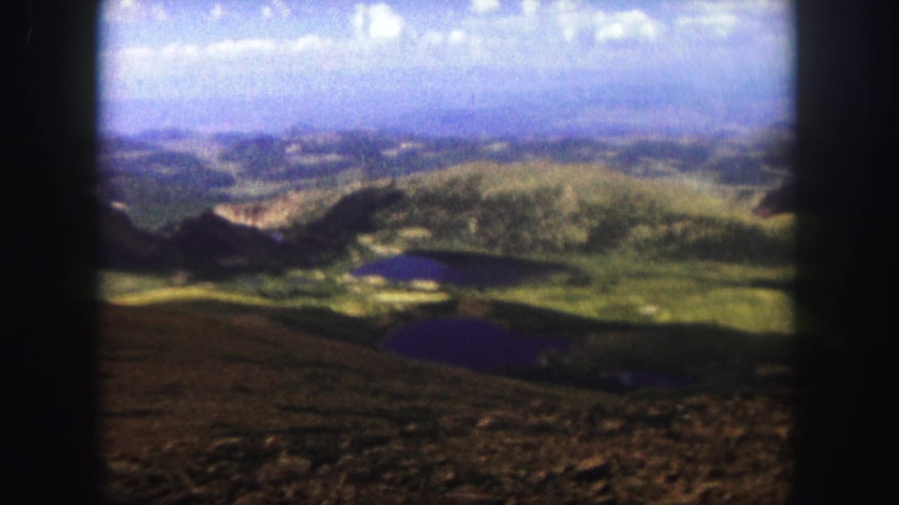 Wide valley panorama with dark lakes below green hills and a soft blue mountain horizon.