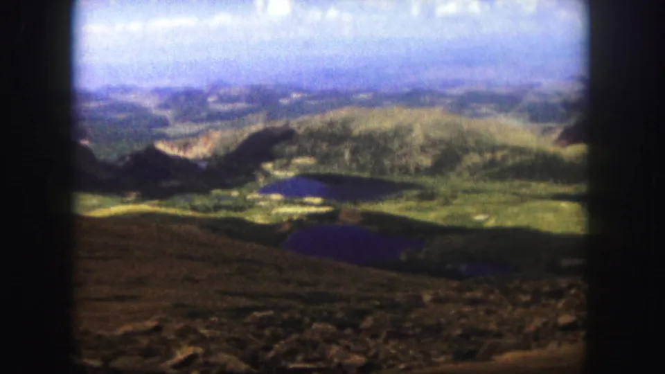 1958 Colorado Springs 8mm still with soft color and grain over a mountain valley, visible lakes, rocky foreground, and distant ridgelines.