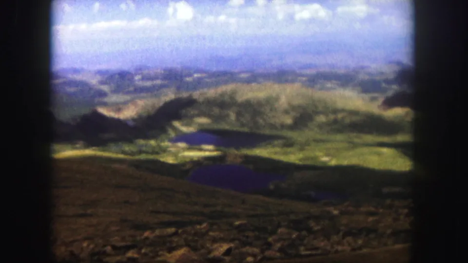 1958 Colorado Springs travel frame showing a mountain overlook toward a lake in a green valley, with rocky foreground and hazy blue distance.