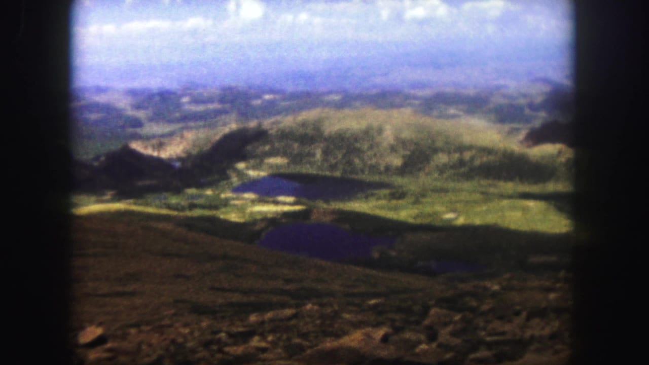 1958 8mm Colorado mountain panorama from Pikes Peak, with green ridges, dark alpine lakes, rocky foreground, and bright sky.