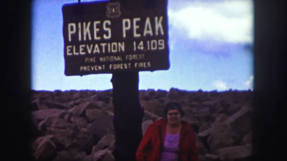 1958 Colorado Springs travel frame with a woman beside the Pikes Peak elevation 14,109 feet sign on rocky summit terrain.