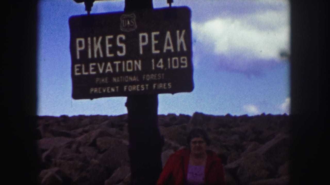 Pikes Peak elevation sign with rocky terrain and a woman standing below.