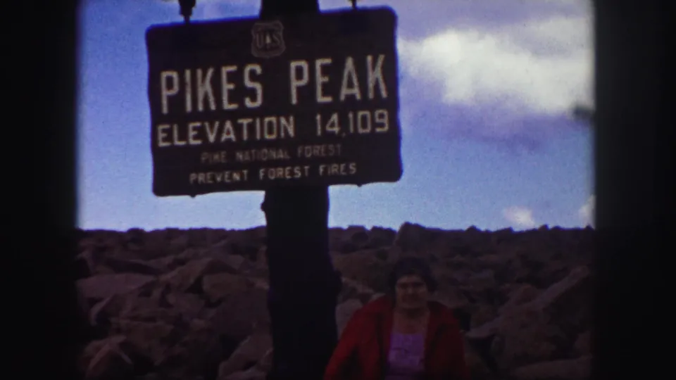 Home-movie frame from Pikes Peak with the elevation sign prominent and a woman in a red jacket near the lower edge.